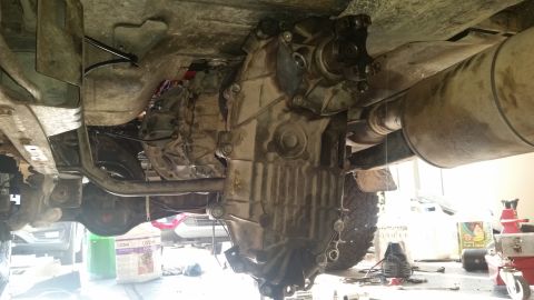 1985 Ford Bronco - Underside view of a vehicle's transmission and transfer case, with suspension components and exhaust visible. Tools and containers are pres…
