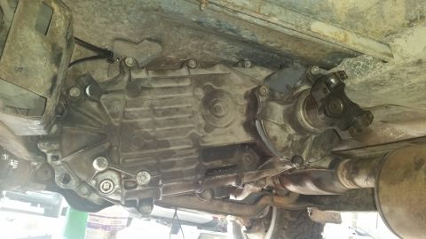 1985 Ford Bronco - The underside of a vehicle shows a transfer case and part of the exhaust system. A driveshaft U-joint is visible connecting to the transfer…