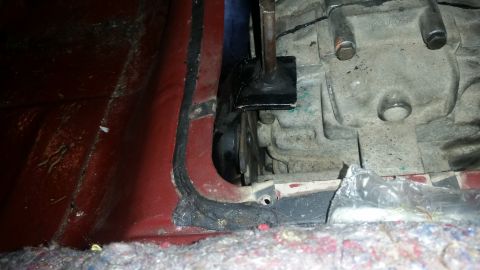 1985 Ford Bronco - A close-up view shows the transmission housing and a black metal bracket attached to it, possibly for a shifter linkage. The vehicle's red…