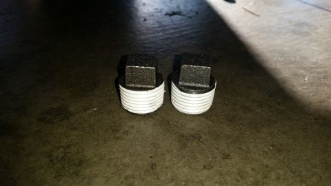 1985 Ford Bronco - Two threaded pipe plugs with square drive heads sit on a dark, textured surface. The plugs appear to be made of metal and have white thread…