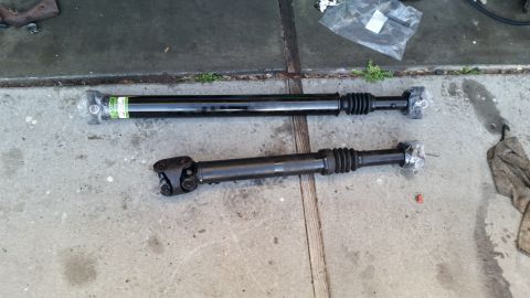 1985 Ford Bronco - Two black driveshafts are laid out on a concrete surface. The top driveshaft is longer and appears new, with a green label and plastic wrap…