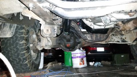 1985 Ford Bronco - Underside view of a vehicle's rear differential and driveshaft, with a large tire visible on the left. A red car is parked behind the vehic…