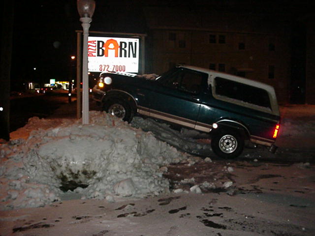 Green Ford Bronco stuck nose-down in a snowbank at night beside a Pizza Barn sign