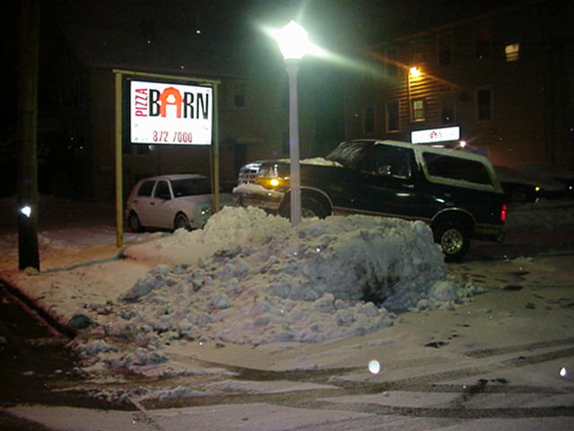 Dark Ford Bronco parked at night behind a snow pile next to a lit Pizza Barn sign