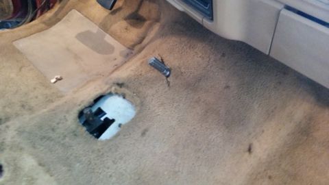 1985 Ford Bronco - Vehicle interior floor showing worn carpet and a hole with exposed mechanical components.
