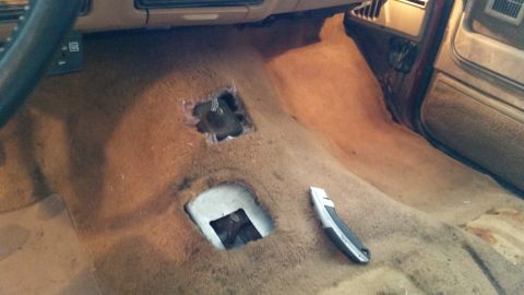 1985 Ford Bronco - The interior of a vehicle shows cut carpet around the transmission tunnel, with a utility knife on the floor.