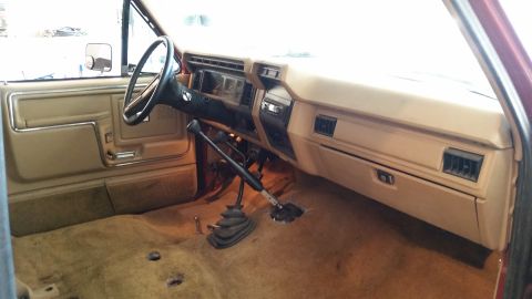 1985 Ford Bronco - The interior of a 1980s Ford Bronco shows a tan dashboard and carpet with a manual transmission.
