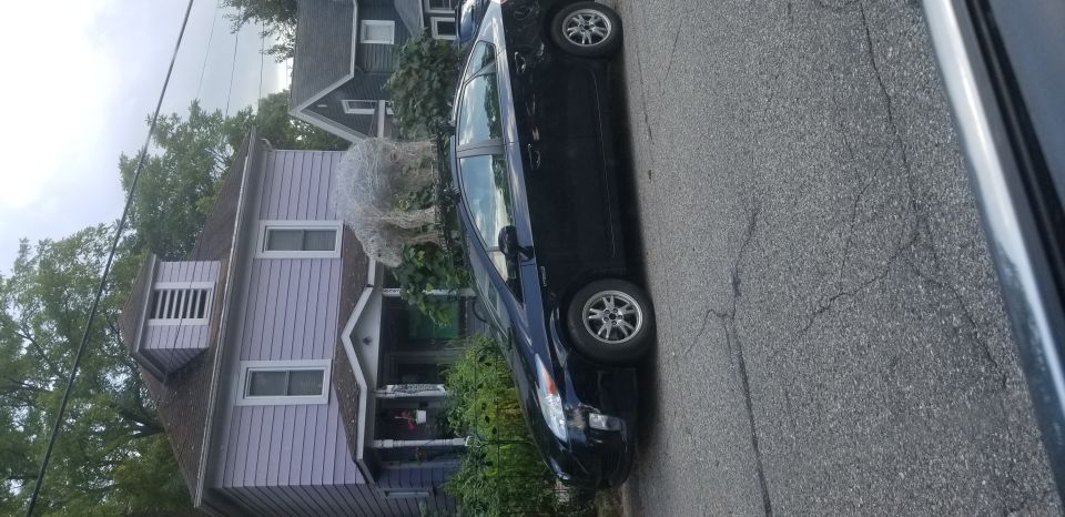 Dark blue sedan parked on cracked asphalt street, side view. A light-colored house with siding is visible behind it