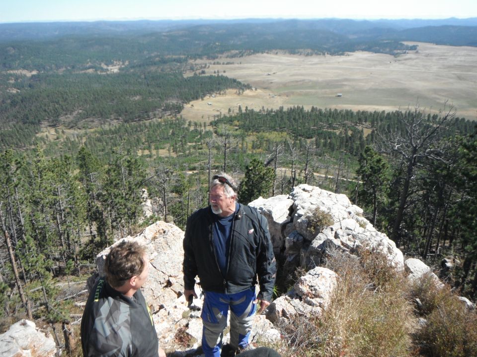 Two men stand on a rocky outcrop overlooking a wide valley with scattered trees and distant hills. A watermark reads SUPERMOTORS