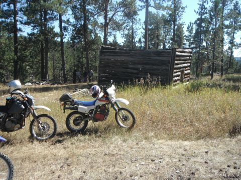 1987 Honda XL600R - Two dirt bikes are parked in a grassy area near a rustic log cabin in a wooded location.