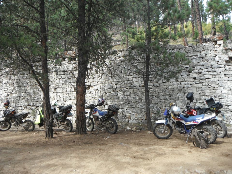 Four dirt bikes parked in front of a rough stone wall with trees around
