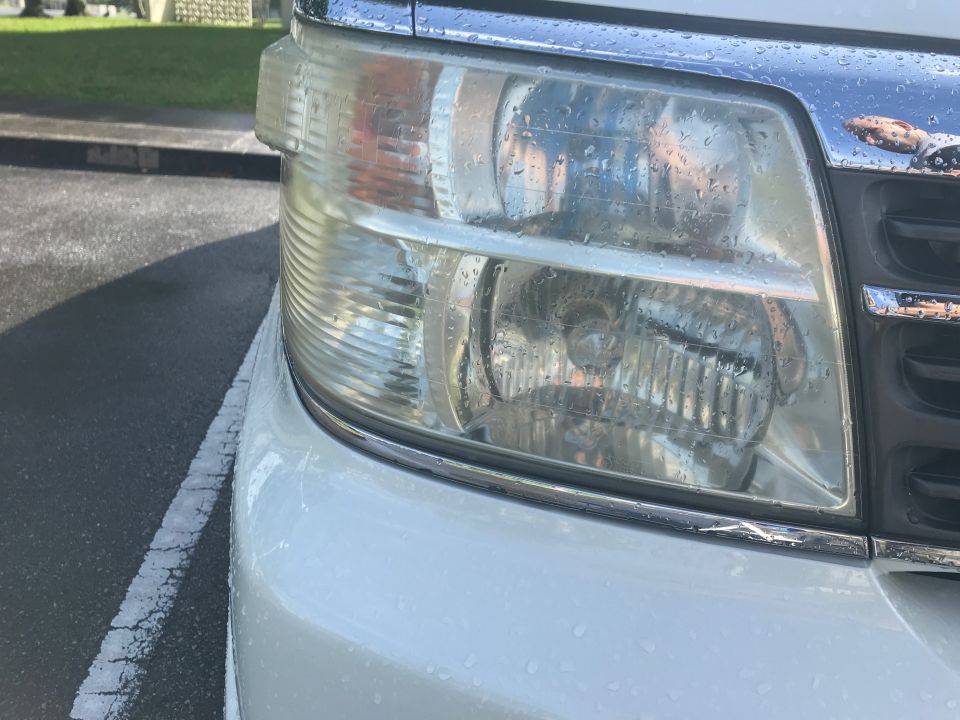 White minivan's front headlight and chrome grille with water droplets. The grille has horizontal bars