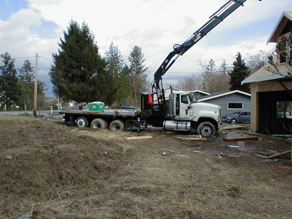 Flatbed truck with crane parked on muddy construction site next to unfinished house. Small car visible in background