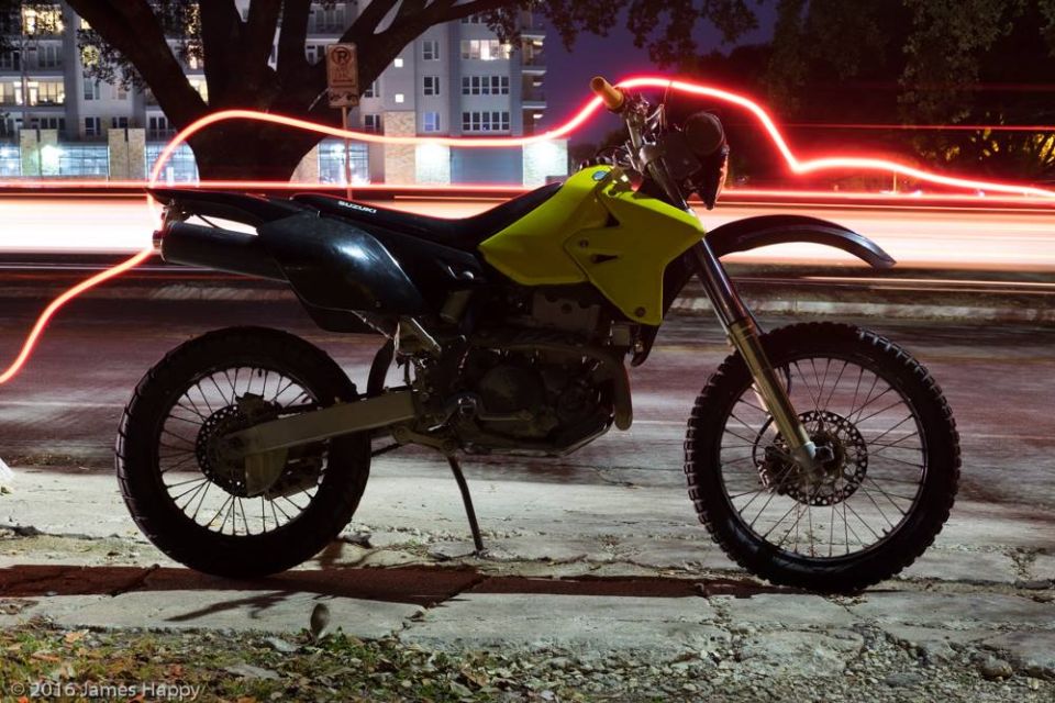 Yellow and black Suzuki dual-sport motorcycle parked at night with red light trails.