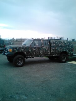 1991 Ford Bronco - A four-door pickup truck with a camouflage paint scheme is parked on a paved surface. The truck has a camper shell with a roof rack and app…