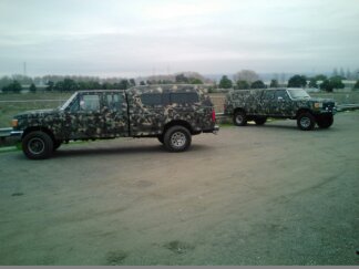 1991 Ford Bronco - Two pickup trucks with camouflage paint are parked on a gravel lot. The truck in the foreground is a four-door pickup with a camper shell,…