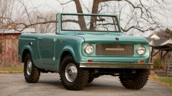 1991 Ford Bronco - A teal, convertible International Harvester Scout 80 is parked on a paved driveway in front of a bare-branched tree and a rustic building.…