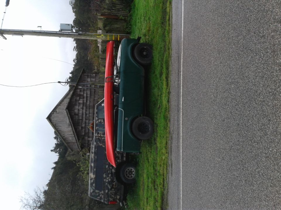 Green vehicle with red kayak on roof parked next to road and building.