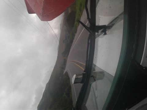 1991 Ford Bronco - View from inside a vehicle looking out the windshield at a wet road winding through a hilly landscape under a cloudy sky. A red object, pos…