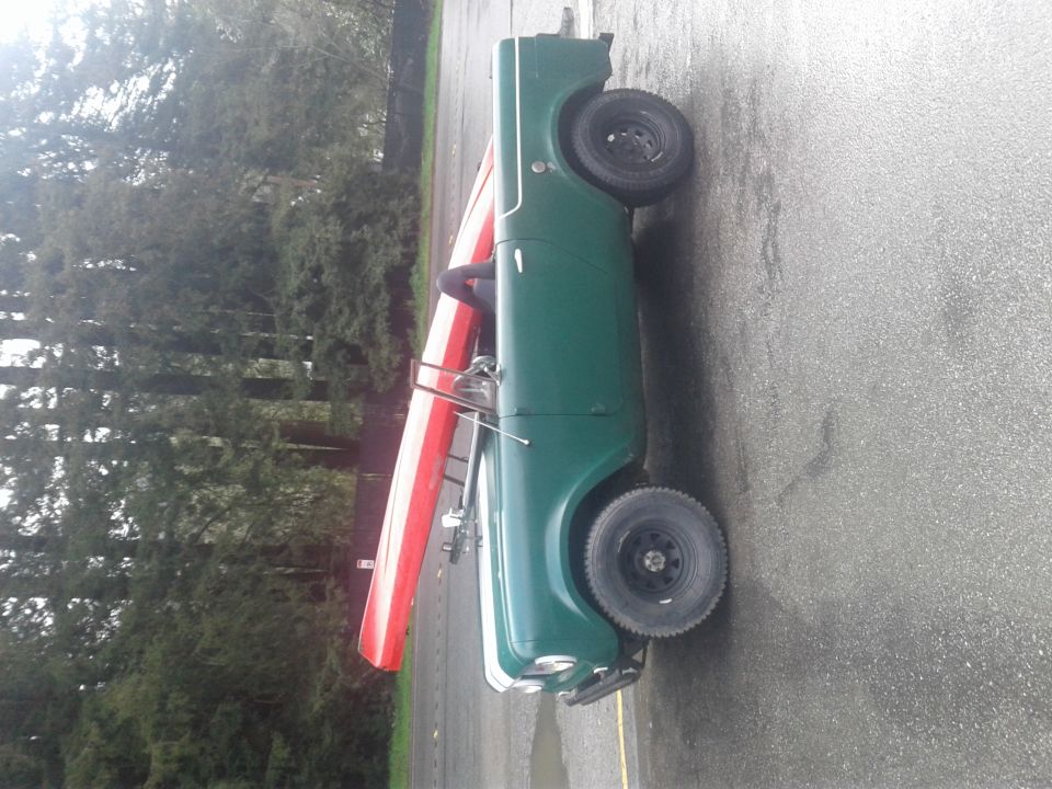 Green vintage Jeep-like vehicle with a canoe on top, parked on asphalt.