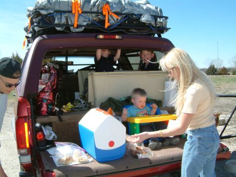 1996 Ford Bronco - A family prepares for a trip, loading a red SUV with gear and children while enjoying snacks.