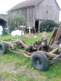 1991 Ford Bronco - The chassis and drivetrain of a 1991 Ford Bronco are shown outdoors in front of a wooden barn. The vehicle appears to be undergoing restora…