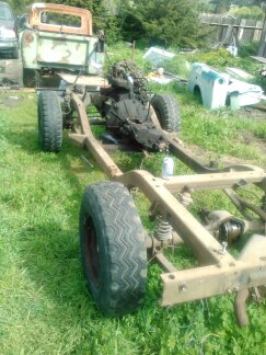 1991 Ford Bronco - The chassis and drivetrain of a vehicle are shown in a grassy outdoor setting. A partial view of a light green pickup truck is visible in t…