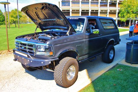1992 Ford Bronco - A lifted dark green Ford Bronco on display at an outdoor car show with its hood propped open and a show card in the windshield reading 'Bra…