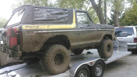 1978 Ford Bronco - A muddy, dark-colored 1978 Ford Bronco with a yellow stripe and "OVERTIME" text is loaded onto a flatbed trailer. The vehicle has a soft to…