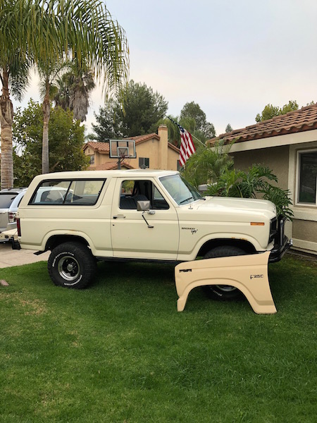 1981 Ford Bronco - A cream-colored 1980s Ford Bronco is parked on a grassy lawn next to a house. A detached fender for a Ford F-250 is lying on the grass in f…