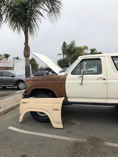 1981 Ford Bronco - A tan fender part is laid on the ground in front of a white and rusty brown Ford Bronco with its hood open. The Bronco is parked outdoors i…