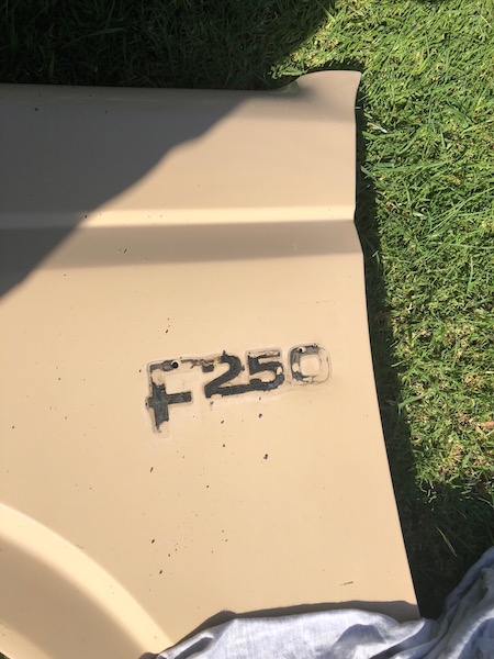 1981 Ford Bronco - A close-up of a tan-colored vehicle panel with the text "F250" embossed on it. The panel is lying on green grass under sunlight.