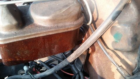 1985 Ford Bronco - A close-up view of a rusty brake master cylinder reservoir with a metal cap. Visible hoses and wiring are present in the engine bay.