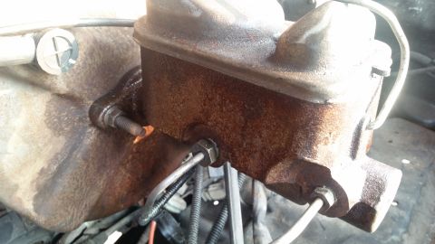 1985 Ford Bronco - A close-up view of a rusty brake master cylinder mounted to a vehicle's firewall. Metal brake lines are connected to the cylinder, with wir…