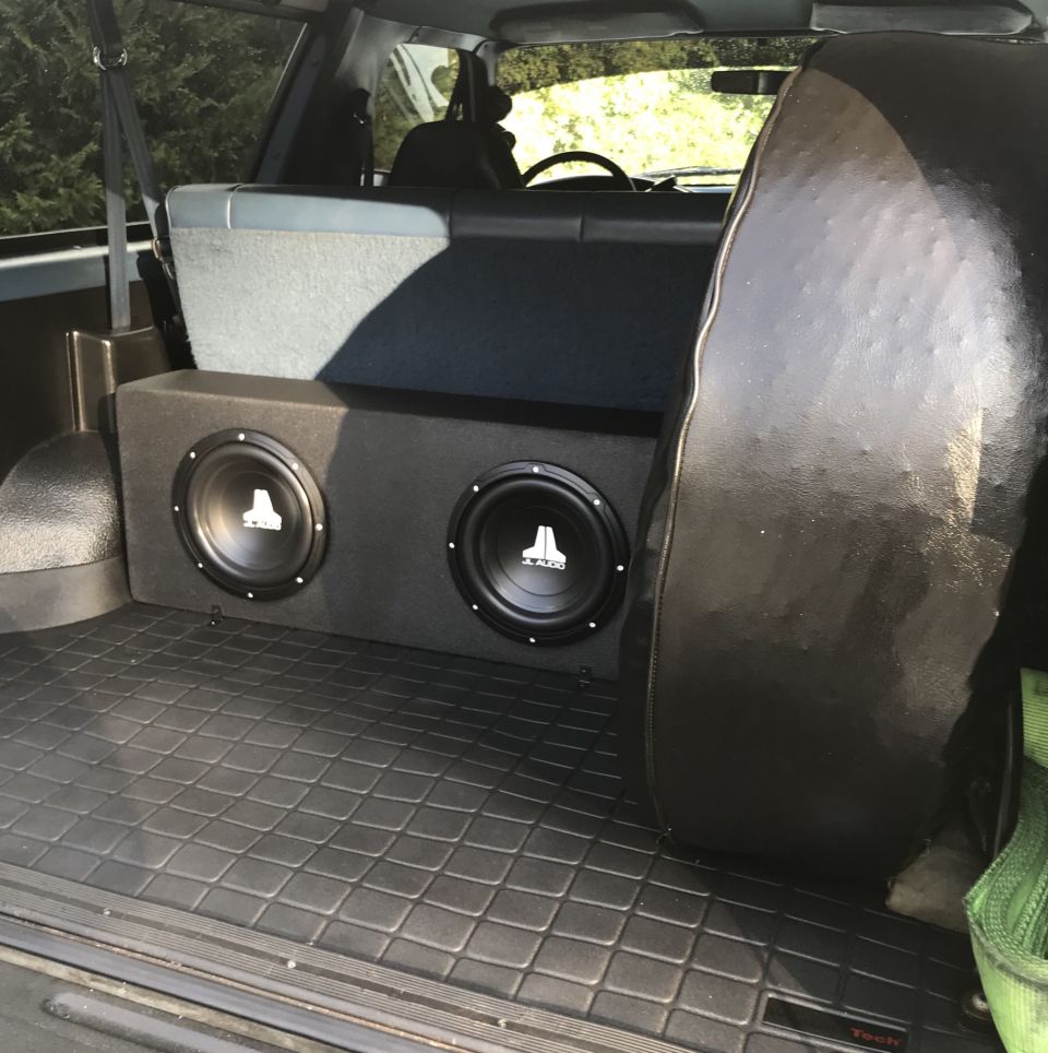Rear cargo area of a vehicle with dual JL Audio subwoofers and a spare tire.