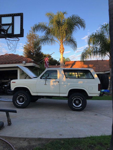 1981 Ford Bronco - A white 1980s Ford Bronco is parked on a concrete driveway in front of a house with a basketball hoop and palm trees in the background. The…