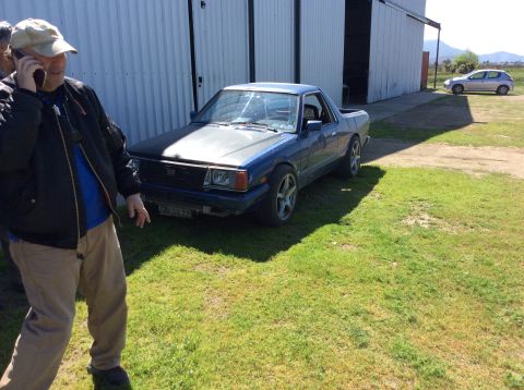 1992 Ford Bronco - A blue Subaru Brat pickup truck is parked on grass in front of a large building with open bay doors. A man wearing a baseball cap and jacke…
