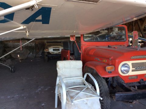 1992 Ford Bronco - A red Toyota Land Cruiser FJ40 is parked under an airplane wing next to a tan FJ40. A white outdoor chair is in the foreground.