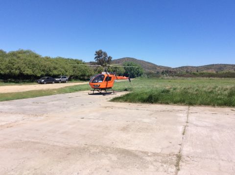 1992 Ford Bronco - An orange helicopter sits on a concrete helipad with a grassy field and hills in the background. Two cars are parked on a dirt path to the…