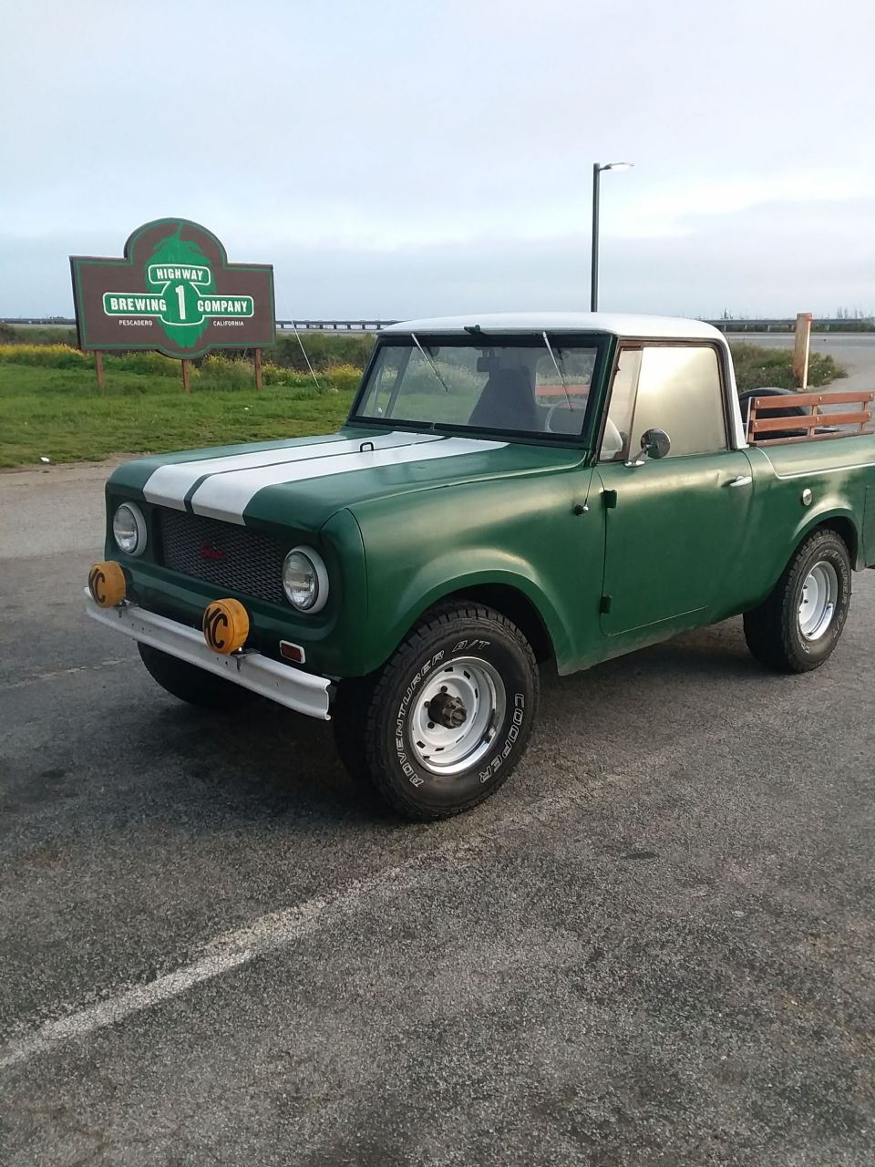Green International Harvester Scout pickup truck with white stripes parked outdoors near a brewery sign.