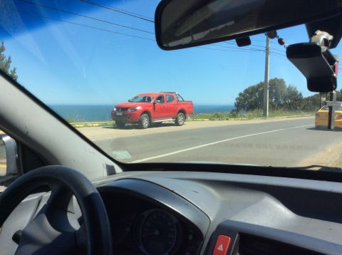 1992 Ford Bronco - A red pickup truck is parked on the side of the road overlooking the ocean, with another vehicle visible in the distance.