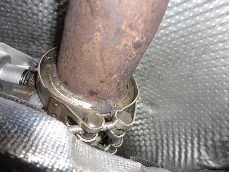 Rusty exhaust pipe with stainless V-band clamp and springs under a car, silver heat shields around it
