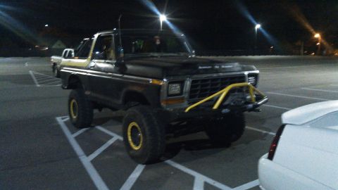 1978 Ford Bronco - A lifted, dark-colored 1978 Ford Bronco with yellow accents on the wheels and a yellow brush guard is parked in a dimly lit parking lot at…