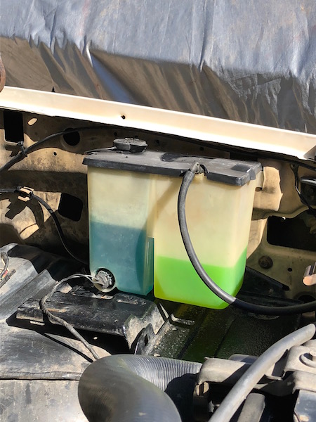 1981 Ford Bronco - A close-up view of a dual-chamber windshield washer fluid reservoir in a vehicle engine bay. One chamber contains blue fluid, and the other…