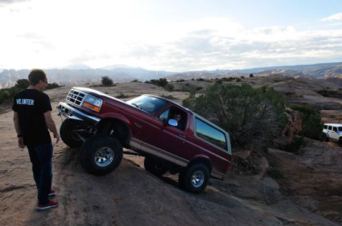 1995 Ford Bronco - A maroon Ford Bronco is navigating a rocky incline with a volunteer standing nearby. Another white SUV is visible in the background.