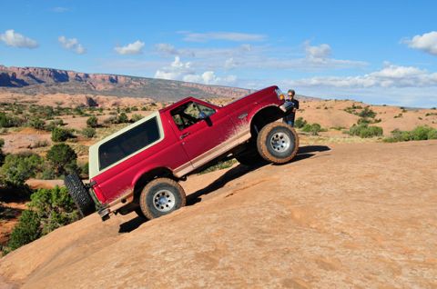 1995 Ford Bronco - A red 1995 Ford Bronco with a white top is angled sharply on a rocky incline in a desert landscape. A person stands by the driver's side do…