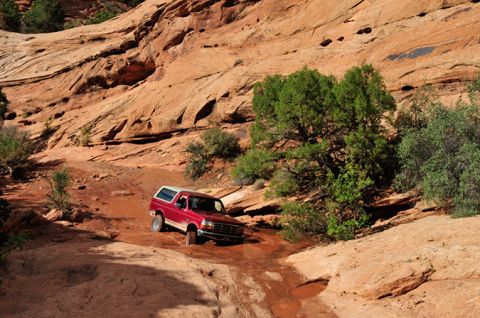 1995 Ford Bronco - A red 1995 Ford Bronco is navigating a rocky, off-road trail with sparse green vegetation and large sandstone cliffs in the background.