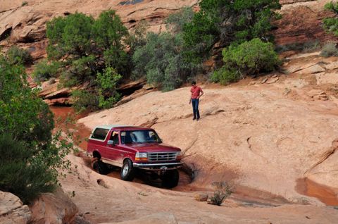 1995 Ford Bronco - A red 1995 Ford Bronco is navigating a rocky, off-road trail with a person standing on a nearby ridge. The vehicle is driving through a dry…