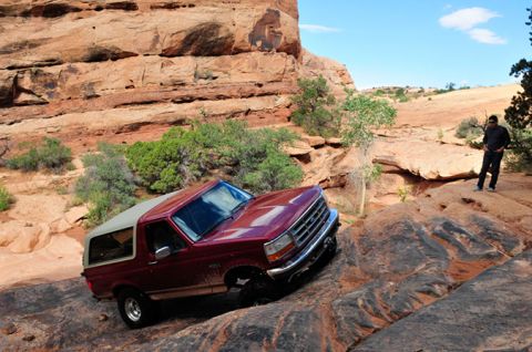 1995 Ford Bronco - A maroon Ford Bronco is navigating a rocky, off-road trail with a person standing on a rock formation in the background. The vehicle is til…