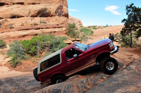 1995 Ford Bronco - A red 1995 Ford Bronco is navigating a rocky, off-road trail with large boulders and red rock formations in the background. A person stands…